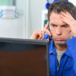 Gestresster Elektriker im Blaumann telefoniert besorgt vor einem Laptop wegen unklarer Mängelhaftung.